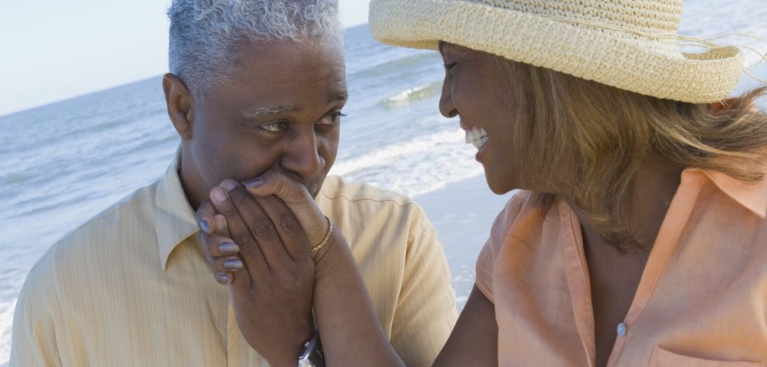 Elderly couple smiling and holding hands on sandy beach, enjoying a peaceful moment together.